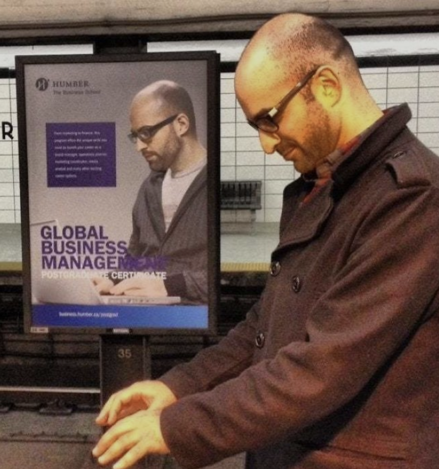 A man standing on a subway platform looks at his phone while wearing a brown coat and glasses. Behind him, an advertisement features a similar-looking man also wearing glasses, promoting a Global Business Management postgraduate certificate.