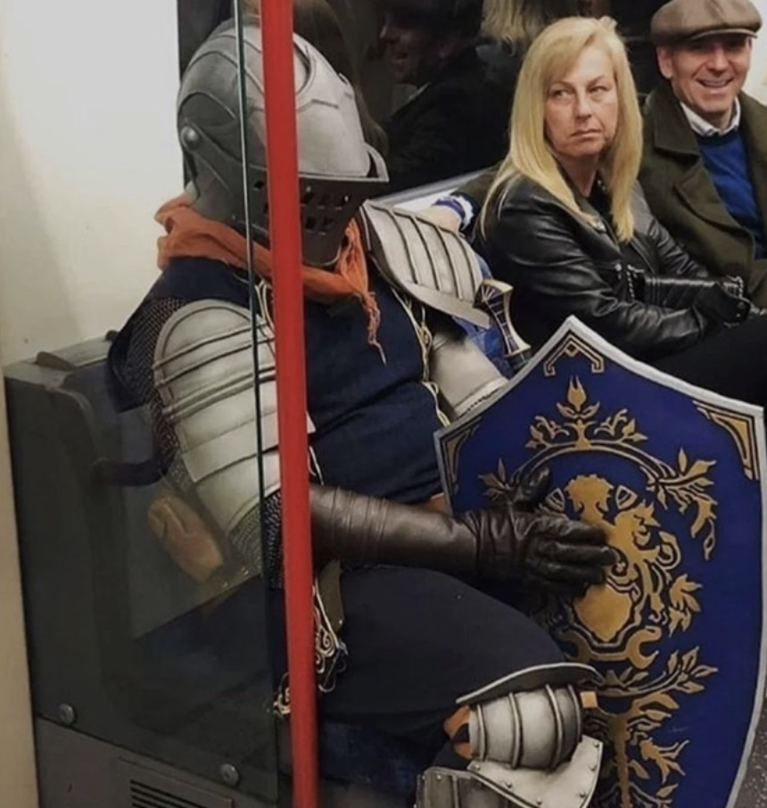 A person wearing full medieval knight armor, complete with a helmet and shield, is sitting on a subway train. Next to them are two casually dressed passengers who appear to be amused and intrigued by the knight's attire.