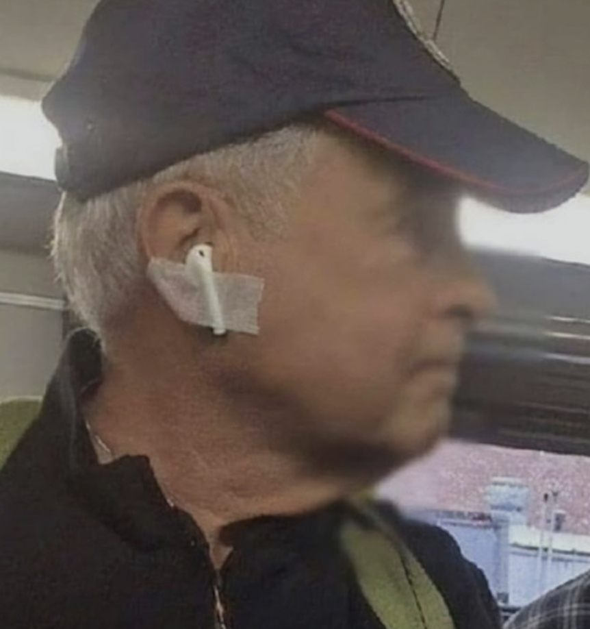 A man with gray hair, wearing a navy blue cap and a black shirt, has an AirPod taped to his ear. He is looking to the right and appears to be on public transportation.