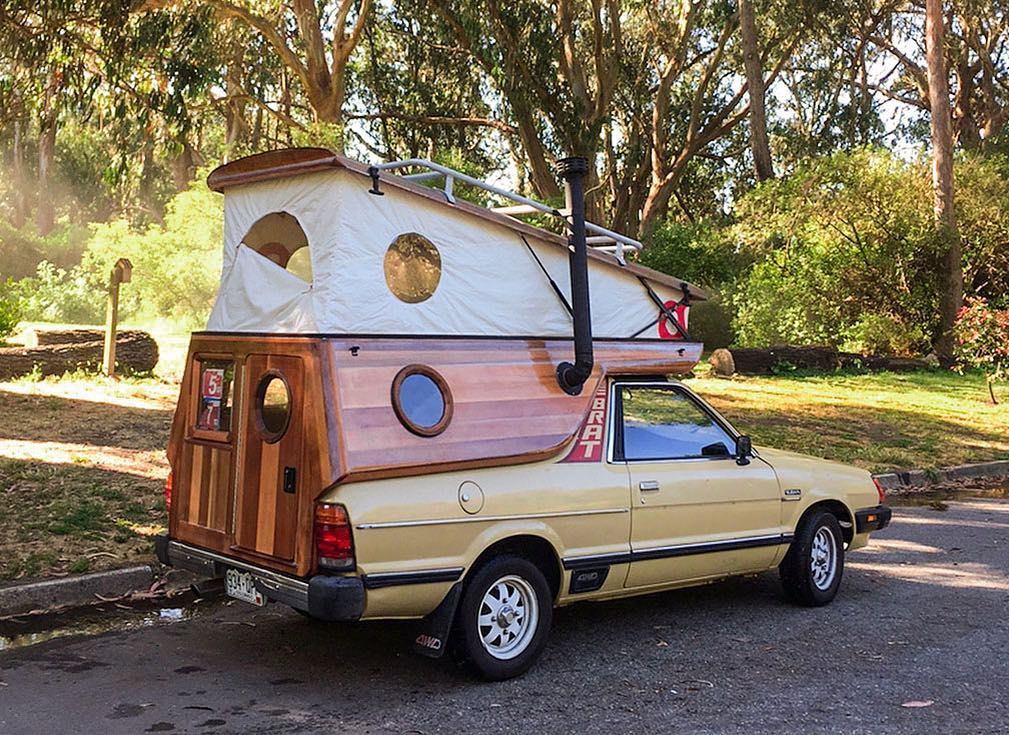 A yellow pick-up truck converted into a camper with a custom wooden cabin structure on the back. The camper features round porthole windows, a canvas pop-top roof, a small chimney, and a bumper sticker that reads "BRT." The vehicle is parked in a wooded area.