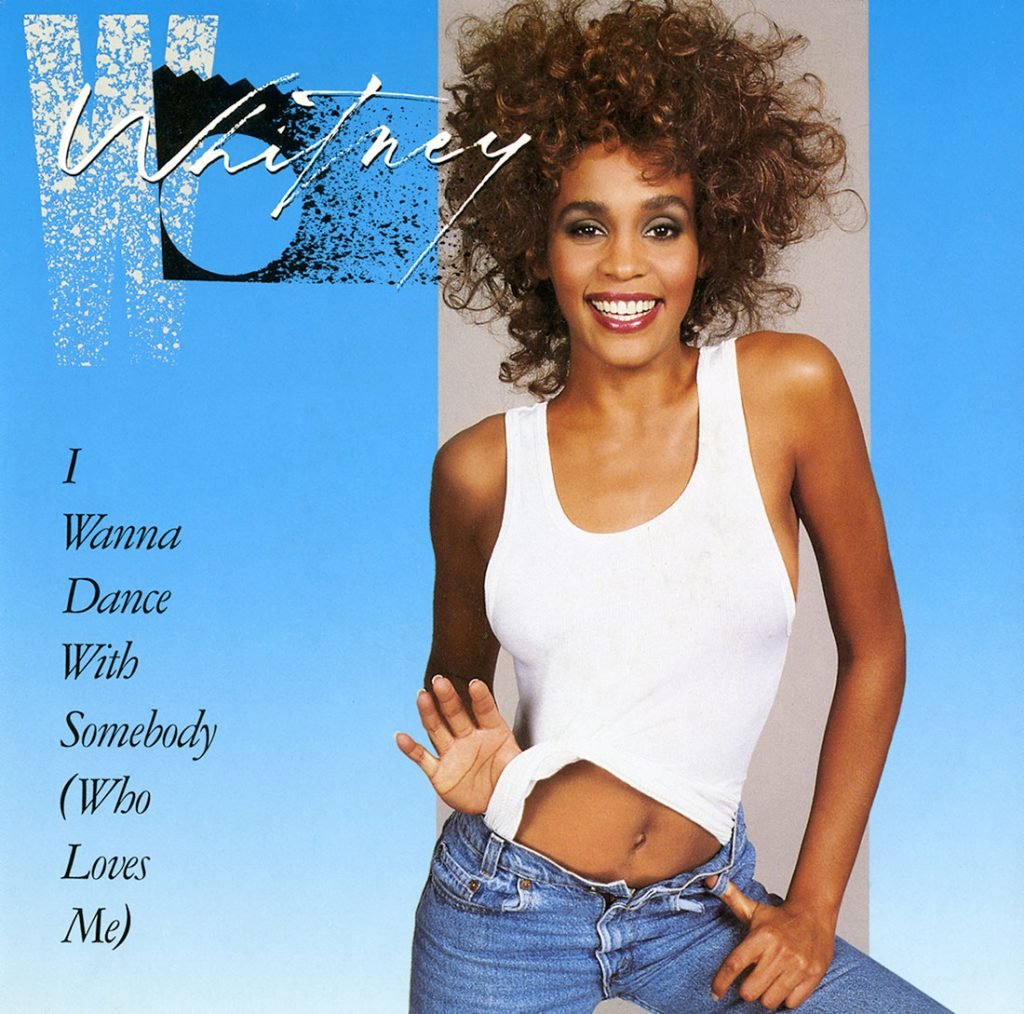 A young woman with curly hair, wearing a white tank top and blue jeans, smiles brightly on the cover for Whitney Houston's single, "I Wanna Dance with Somebody (Who Loves Me)." The background is blue, fading to white with the text in black on the left side.
