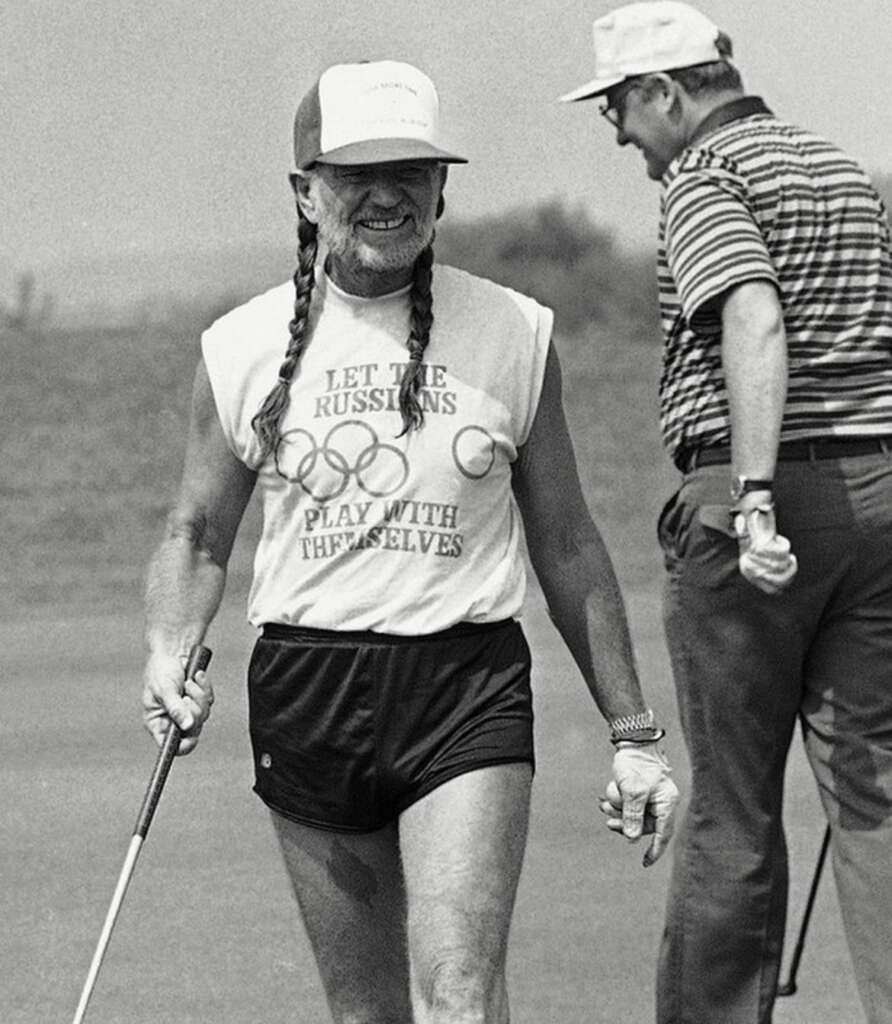 A smiling person with long braids and a trucker hat is holding a golf club on a golf course. They are wearing a sleeveless shirt with text and an Olympic rings graphic, and short shorts. Another person in a striped shirt and cap is in the background, looking away.