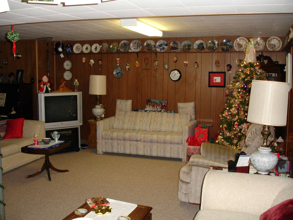A cozy living room with wood-paneled walls is decorated for Christmas. A lit Christmas tree stands in the corner, surrounded by presents. The room features a vintage TV, a striped couch, various wall plates and decor, and several lamps providing a warm ambiance.
