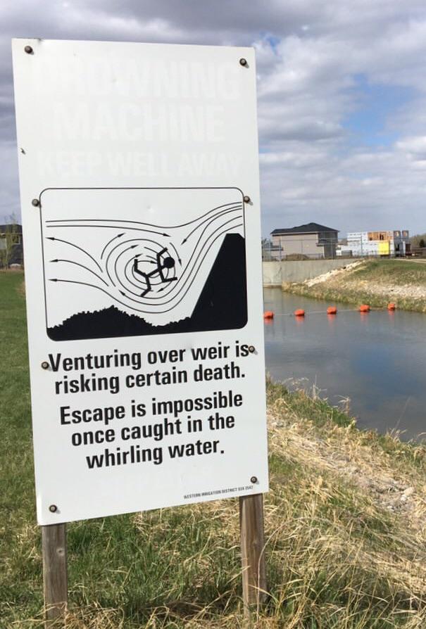 A warning sign near a waterway reads: "Venturing over weir is risking certain death. Escape is impossible once caught in the whirling water." The sign features an illustration of a person being swept into dangerous, swirling water near a weir.