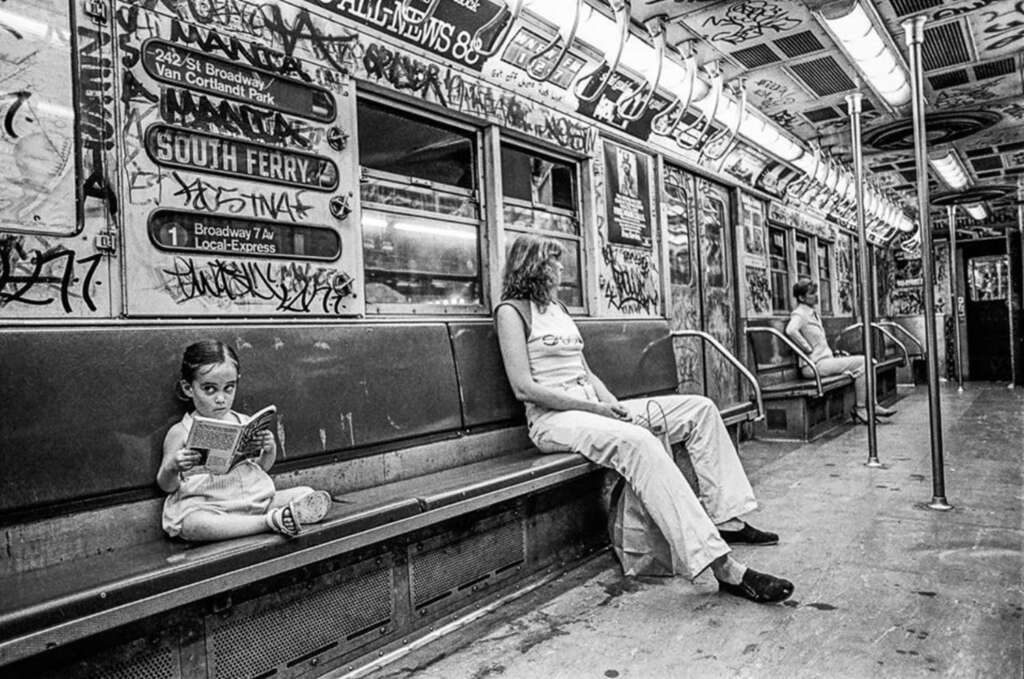 Black and white photo of a graffiti-covered subway car. A young girl sits reading a book on the bench, while a woman sits nearby. Two other passengers are seated on the opposite side. Numerous handwritten notes and tags cover the walls and roof of the car.