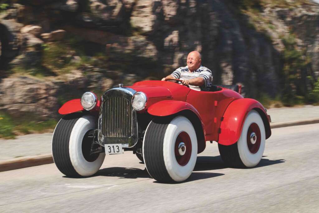 A man drives a unique, vintage-style red car with large white-walled tires on a sunny day. The road is bordered by rocky terrain, and the man's expression conveys enjoyment. The car has the number "313" on the front.