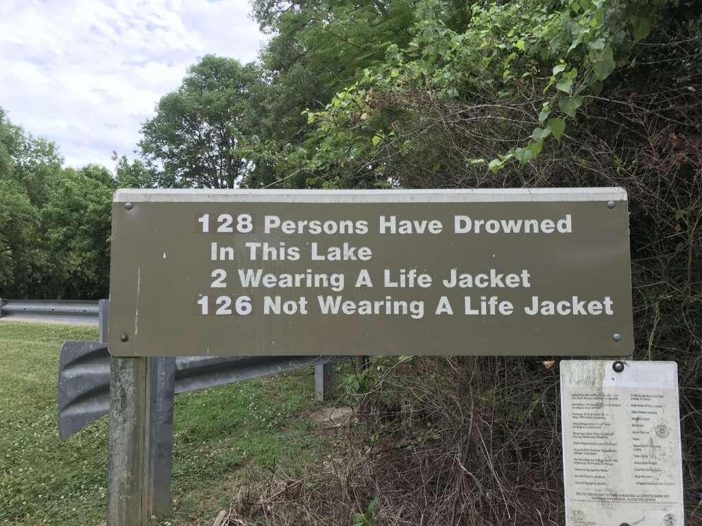 A brown sign near a lake surrounded by trees reads: "128 Persons Have Drowned In This Lake, 2 Wearing A Life Jacket, 126 Not Wearing A Life Jacket." The sign emphasizes the importance of wearing life jackets. A smaller, partially visible sign is posted beneath it.