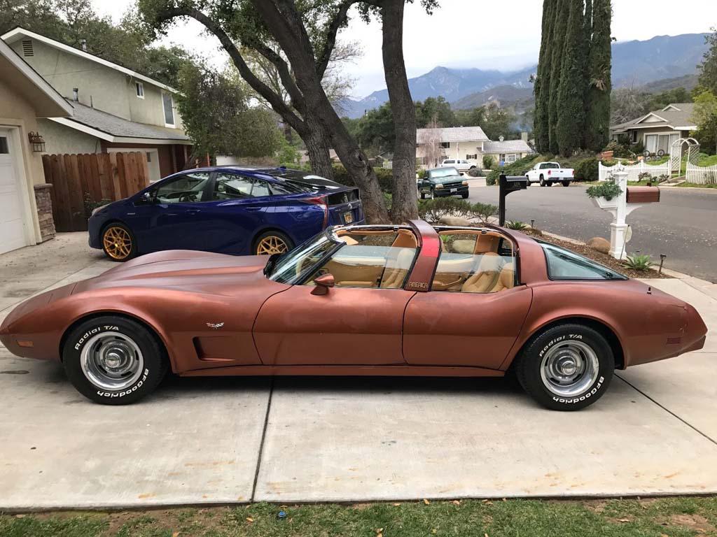 A vintage brown sports car with a T-top roof is parked in a residential driveway. It has "Corvette" written on the side and white-letter tires. In the background, a blue car with gold rims is parked, and suburban houses and trees are visible.