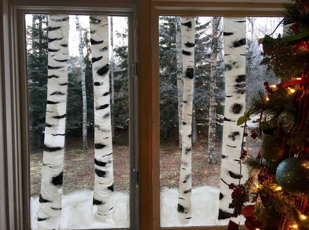 A view through two windows with a painting of white birch trees on the glass panes. Outside, a wooded area is visible. On the right edge, part of a decorated Christmas tree with ornaments and lights is seen.