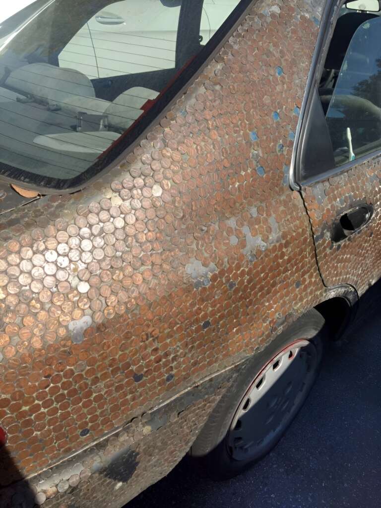 A car partially covered in pennies is parked in a lot. The close-up view showcases a rear door and fender meticulously adorned with coins, creating a unique and shiny mosaic effect. The car's window and part of the interior are also visible.