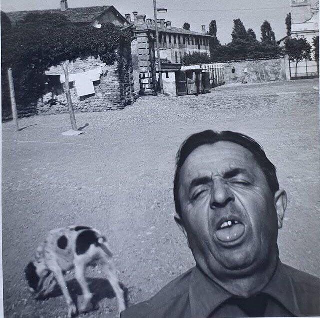 Black and white photo of a man with eyes closed and mouth open, standing in a dirt area. Behind him, there is a dog sniffing the ground, clothes hanging on a line, old buildings, and trees in the distance. The image has a candid, humorous feel.