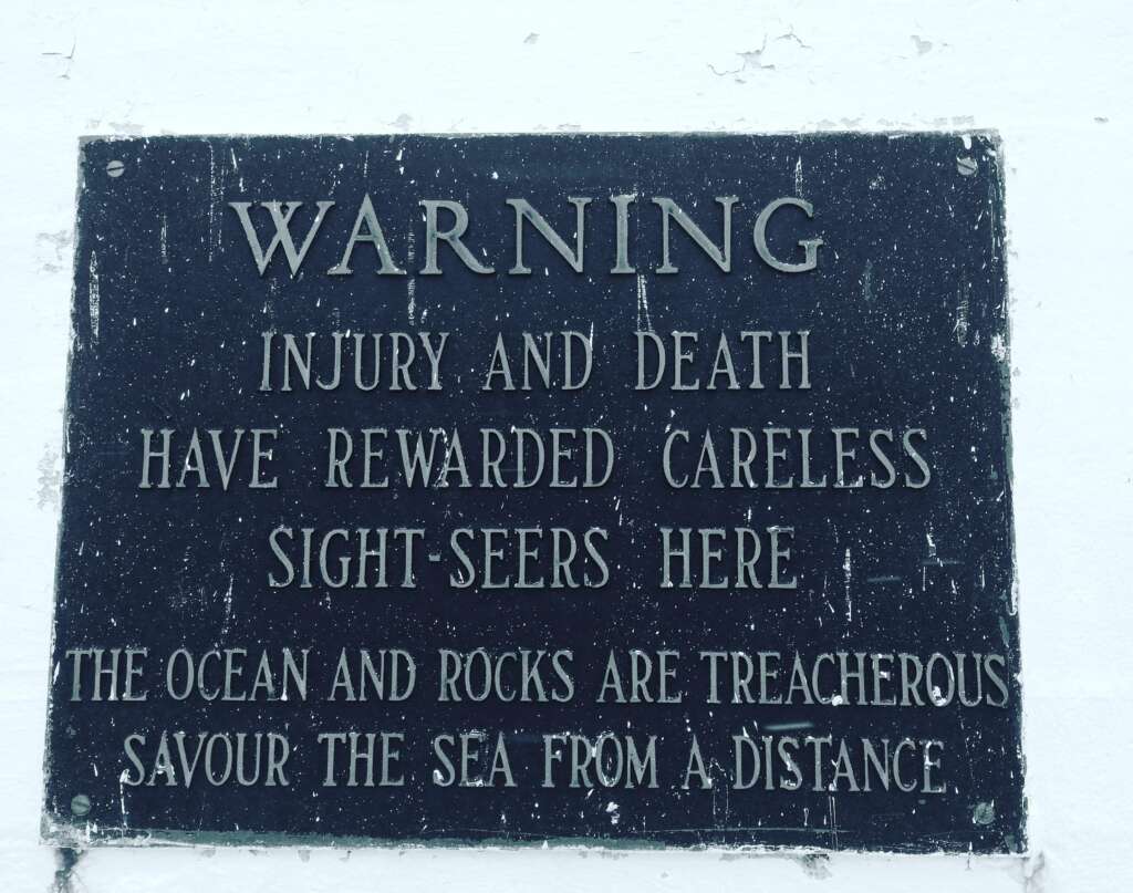 A weathered sign with white text on a black background reads: “WARNING. Injury and death have rewarded careless sightseers here. The ocean and rocks are treacherous. Savour the sea from a distance.” The sign is mounted on a white wall.
