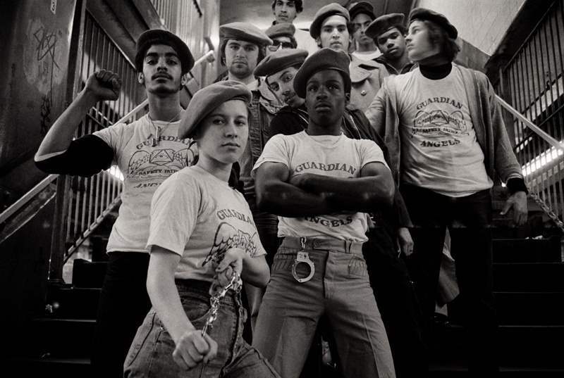 A group of people wearing matching "Guardian Angels" shirts and berets stand on a staircase. They exhibit determined and confident poses, with some crossing their arms and one in front raising his fist. The setting appears to be an urban environment.