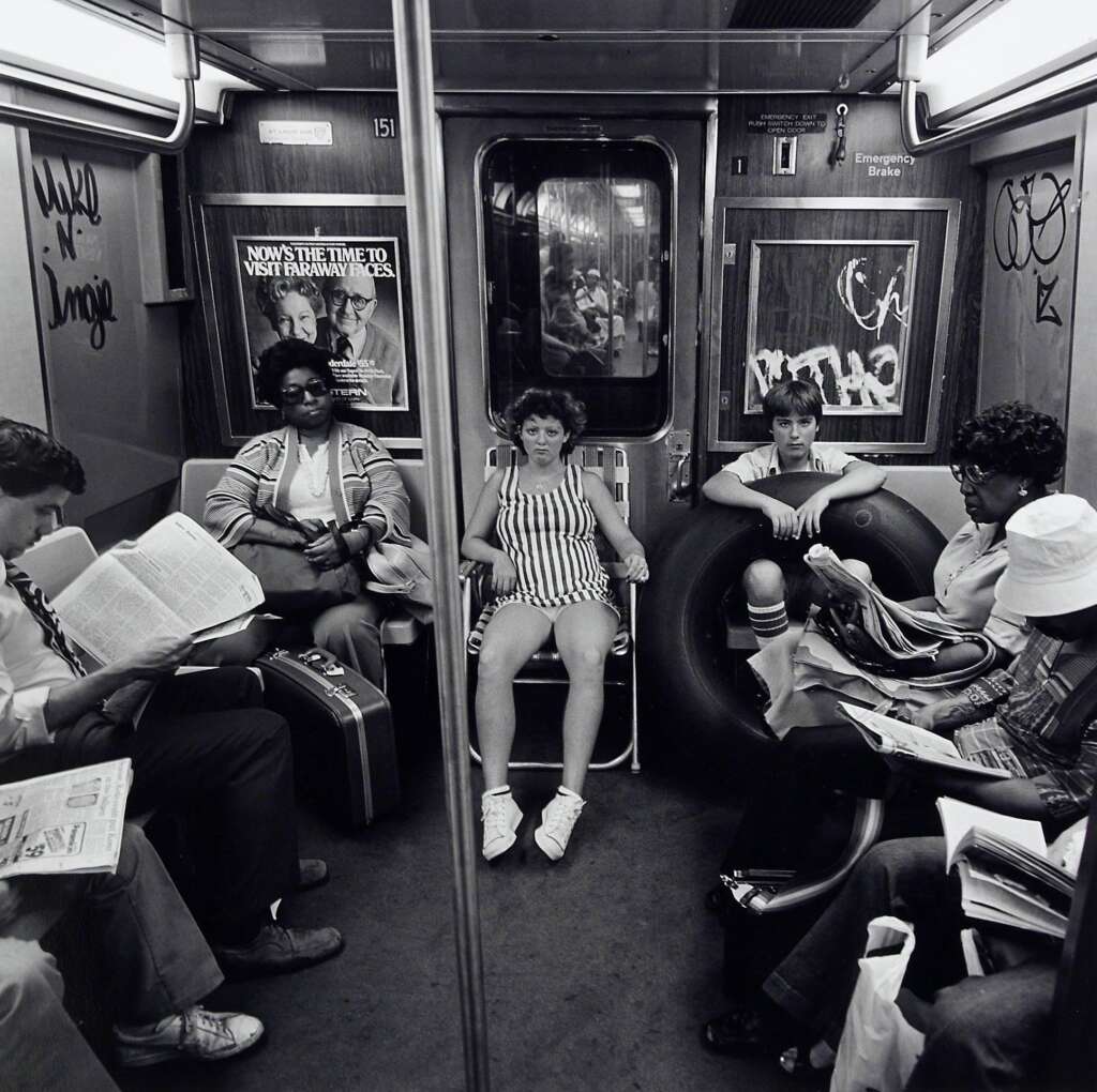 A black and white photo of people sitting on a train. The passengers include a person reading a book, a person in a striped outfit sitting on a folding chair, and others engaging in various activities. The walls are graffitied, and a tourism advertisement is visible.