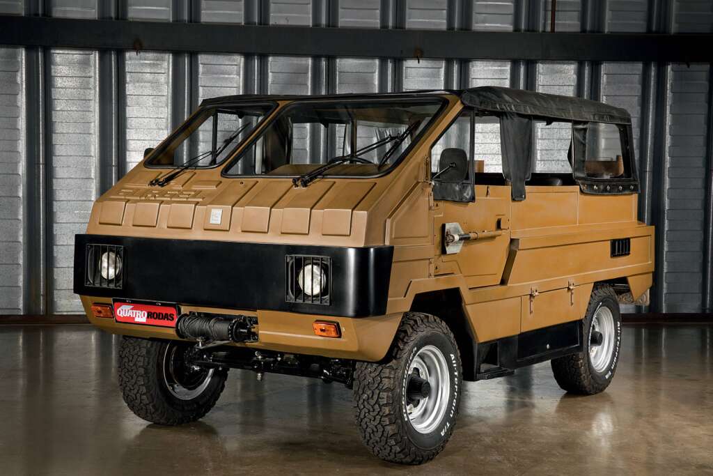 A rugged, tan-colored off-road vehicle with a boxy design is parked indoors on a concrete floor. It features large tires, distinctive front grilles with square headlights, and a soft top. The vehicle appears to be well-suited for rough terrains.