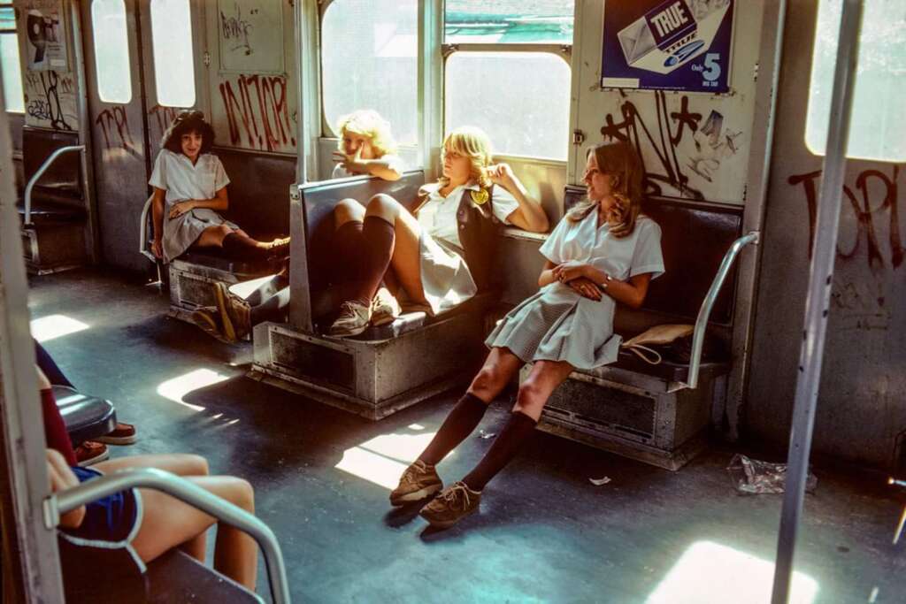 Four young women in school uniforms sit relaxed on a graffiti-covered subway train. Two sit together on a bench on the right side, one lounges with her legs across a seat, and another sits on the left side. Daylight filters through the windows.