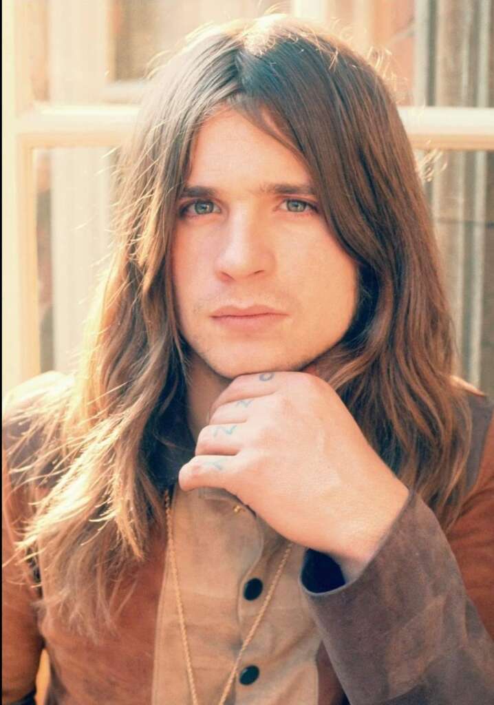 A person with long brown hair and blue eyes is posing for a photo with their hand resting on their chin. They are wearing a light brown button-up shirt and several necklaces. The background is softly lit with natural light coming through a window.