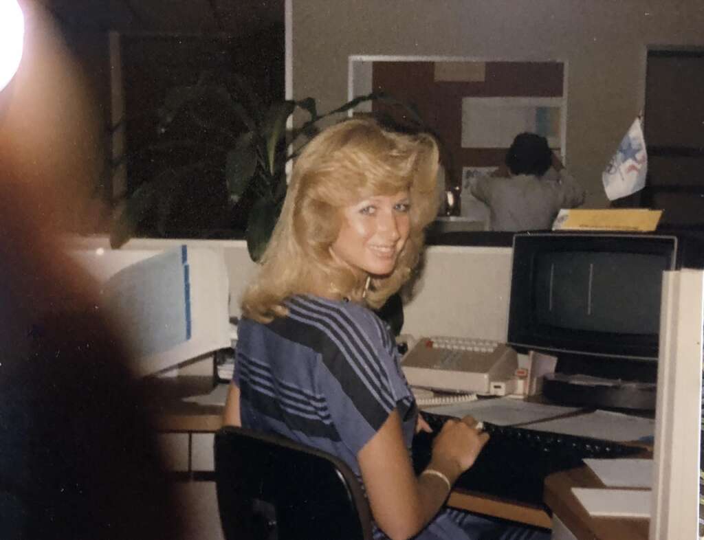 Smiling woman with blonde hair sits at a desk in an office, wearing a blue-striped blouse. An old computer monitor and a typewriter are on her desk. A person with back turned is visible in the background. The setting appears to be from the 1980s.