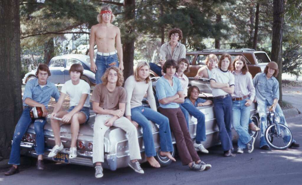 A group of young people, mostly men, pose casually with arms crossed or leaning on a white convertible car parked outdoors. Some are sitting on the hood, and there's a mini bike beside the car. They all wear casual 1970s attire, including jeans, t-shirts, and headbands.