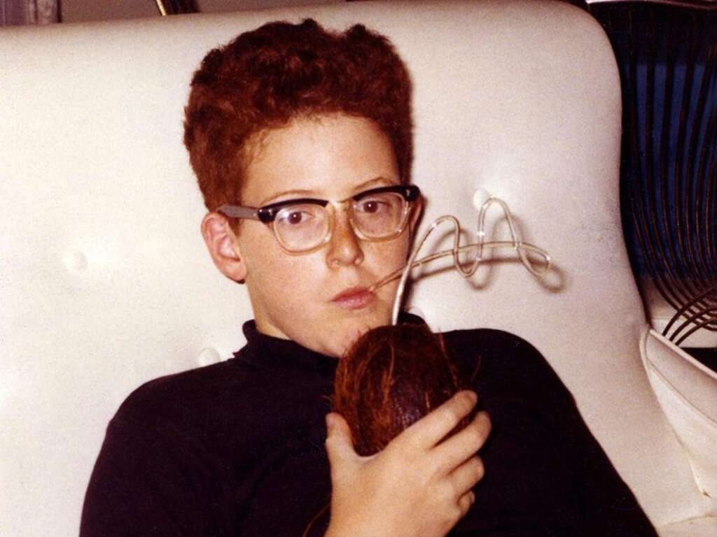 A young person with curly red hair and glasses is relaxing on a white couch, sipping a drink through a curly straw inserted into a coconut. The person is wearing a dark shirt and appears to be deep in thought or simply enjoying the drink.