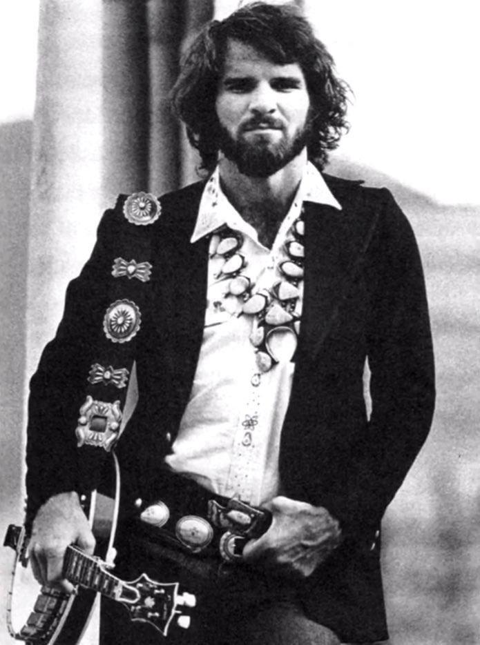 A man with long hair and a beard is wearing a black jacket adorned with large, ornamental conchos and a decorative belt. He is holding a banjo. The photo is in black and white.