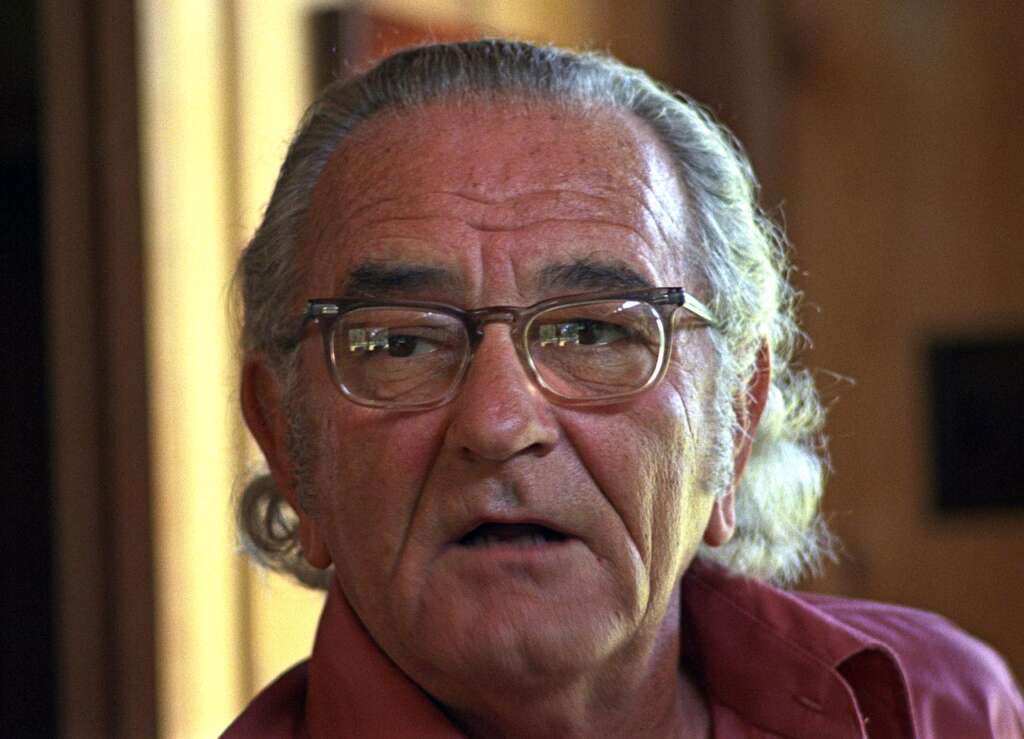 An elderly man with grey hair and glasses is wearing a red shirt. He appears to be speaking or reacting to something, with his mouth slightly open and a serious expression on his face. The background is out of focus.