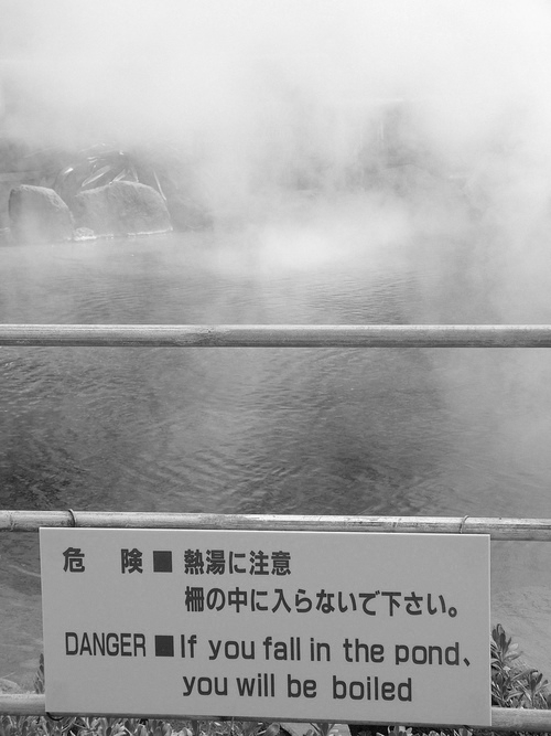 A pond surrounded by mist has a sign attached to a railing in front of it. The sign contains text in Japanese and English warning: "DANGER - If you fall in the pond, you will be boiled." Some greenery is visible at the bottom.