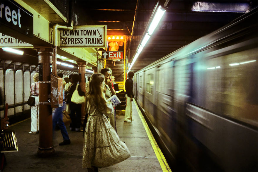 A woman in a long dress stands on a subway platform holding a magazine and looking towards an approaching train. Another person stands nearby, with a sign above indicating "DOWNTOWN EXPRESS TRAINS" and an exit sign. The train is partially blurred in motion.