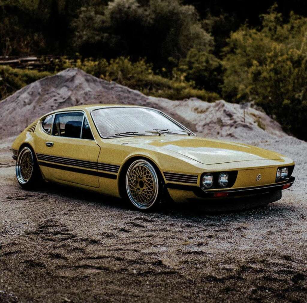 A vintage yellow sports car with sleek, rounded edges and thin, black racing stripes parked on a gravel surface. The car features large, gold rimmed wheels and a low, aerodynamic profile. Trees and a small hill are visible in the background.