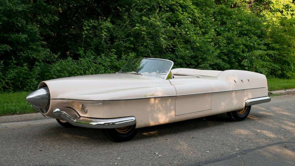 A vintage, cream-colored convertible car is parked on a street with lush green trees in the background. The car features a distinctive, pointed front end and a sleek, streamlined design. The roof is down, and bright sunlight illuminates the scene.