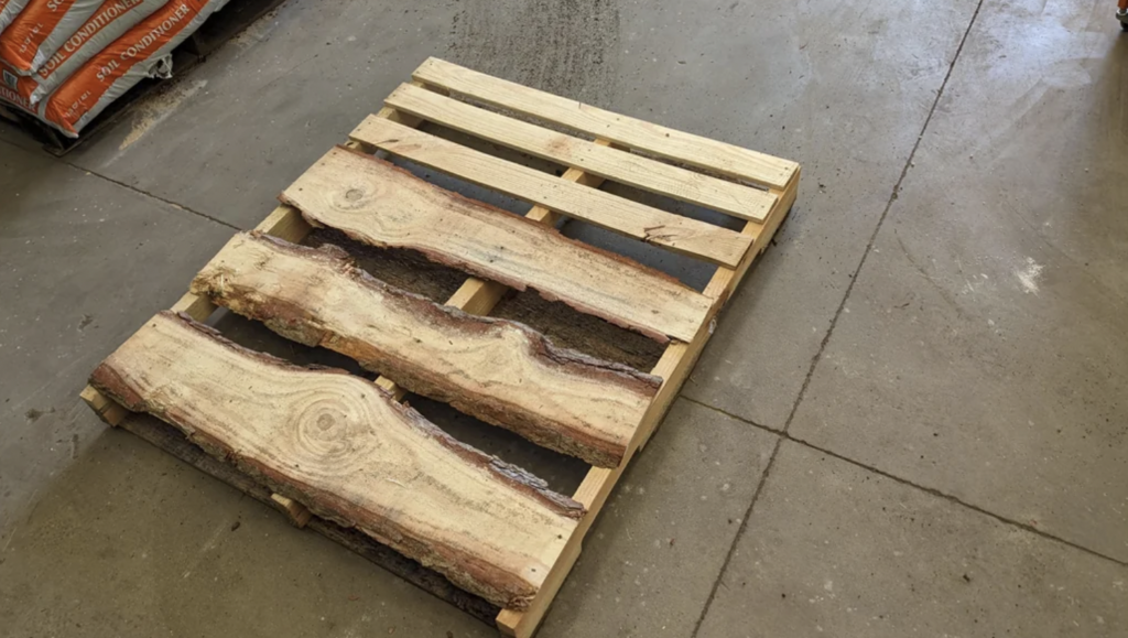 A wooden pallet with rough, unfinished planks placed on a gray concrete floor. The top planks have a natural, rugged texture with visible bark and irregular shapes. Some bags of materials are visible in the background to the left.