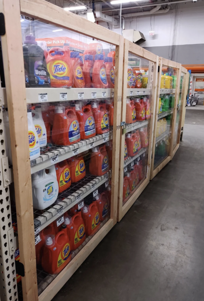 Shelves stocked with large bottles of laundry detergent encased in wooden and clear plastic barriers for security. Various brands and colors are visible, including Tide, All, and Woolite, in a warehouse store setting with concrete floors and industrial lighting.