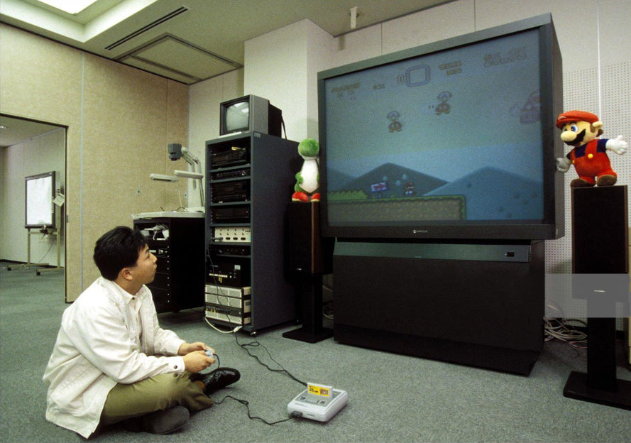 A person sits on the floor, holding a game controller, facing a large TV screen displaying a Super Mario game. Several electronics and stuffed toys, including Mario and Yoshi, are beside the TV in a room with office furniture and equipment.