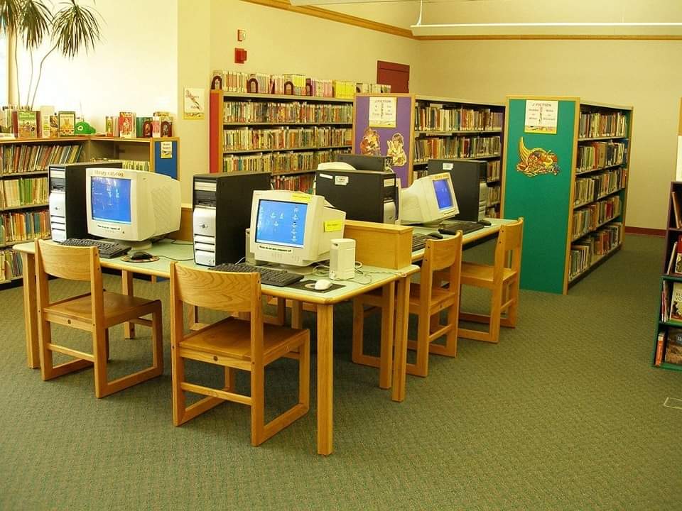 Interior of a library showcasing rows of bookshelves filled with books. In the center are four wooden workstations with beige computers and monitors on them. Wooden chairs are placed at each workstation, and a potted plant is visible in the background.