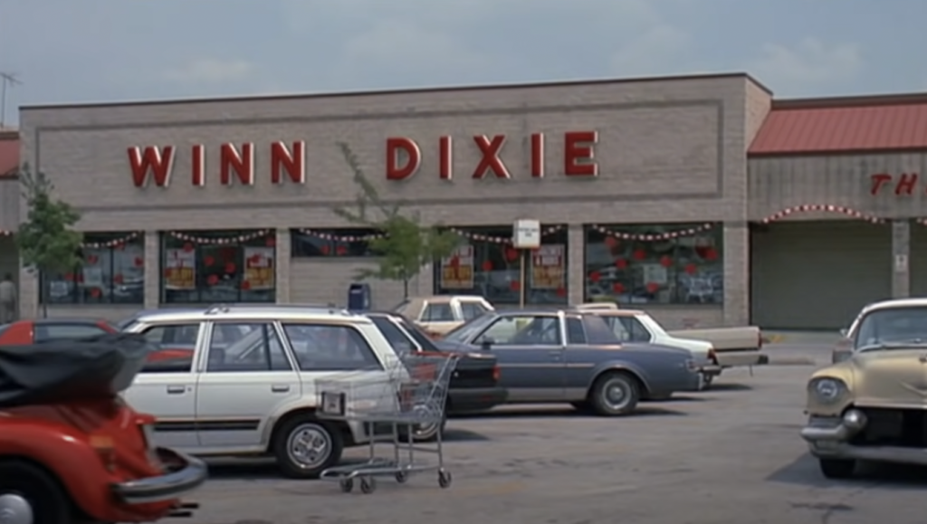 A supermarket with the sign "WINN DIXIE" in red letters on the building facade. The parking lot in front has several cars and a shopping cart. The day appears to be clear with a light blue sky and some trees in the background.