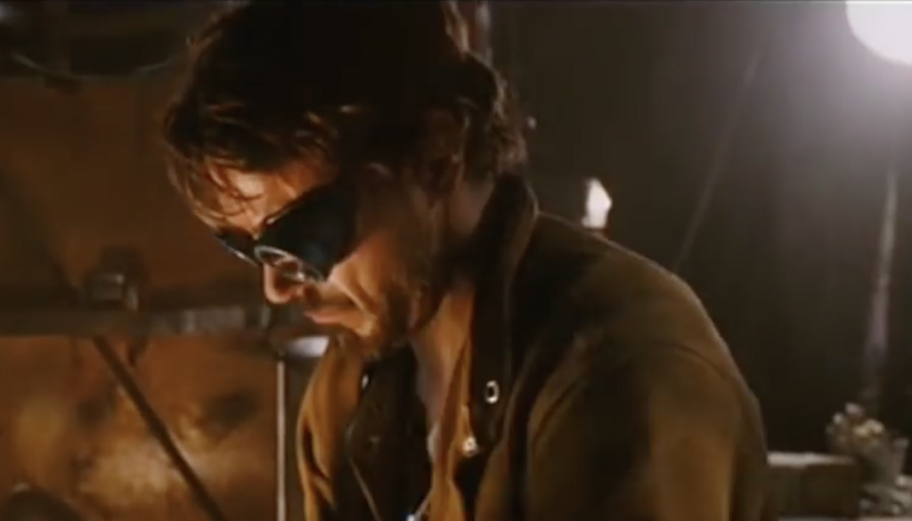 A man with messy hair wearing dark safety goggles and a brown jacket is focused on his work. He is in a dimly lit industrial setting with a bright light in the background, suggesting he might be working on something mechanical or technical.