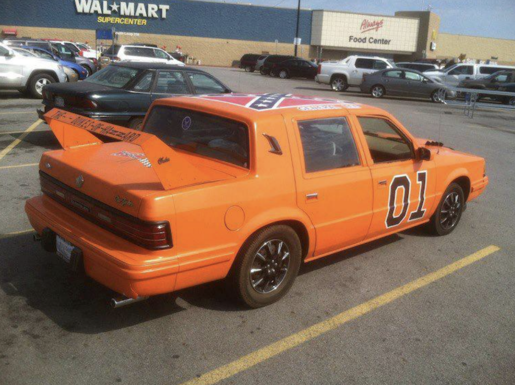 An orange car modified to resemble the "General Lee" from "The Dukes of Hazzard," complete with the Confederate flag on the roof and the number 01 on the side, is parked outside a Walmart Supercenter. The car has black rims and other custom details.