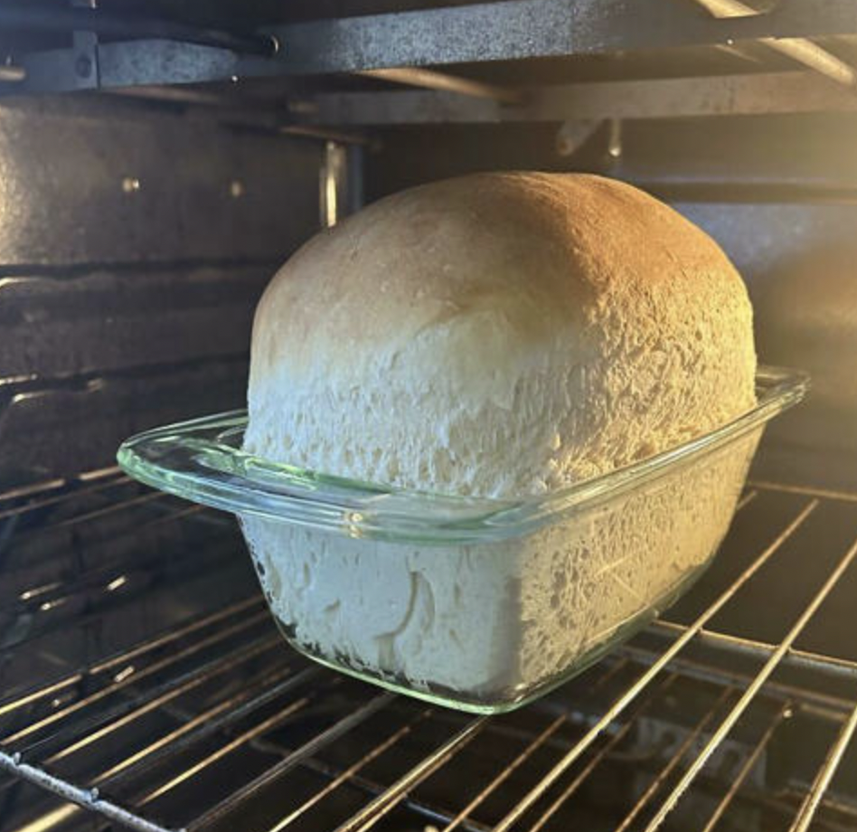 A golden-brown loaf of bread is baking in a glass loaf pan inside an oven. The bread has risen to a rounded top and appears soft and fluffy. The oven interior is lit, highlighting the warmth and texture of the freshly baked bread.