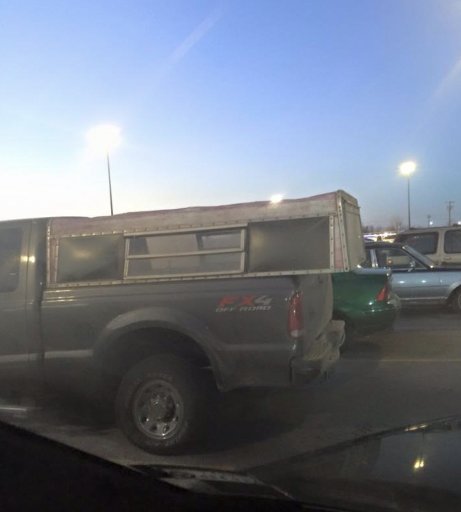 A grey pickup truck with a camper shell is parked in a parking lot during early evening. Overhead lights illuminate the area, and other vehicles are visible in the background under a clear sky.