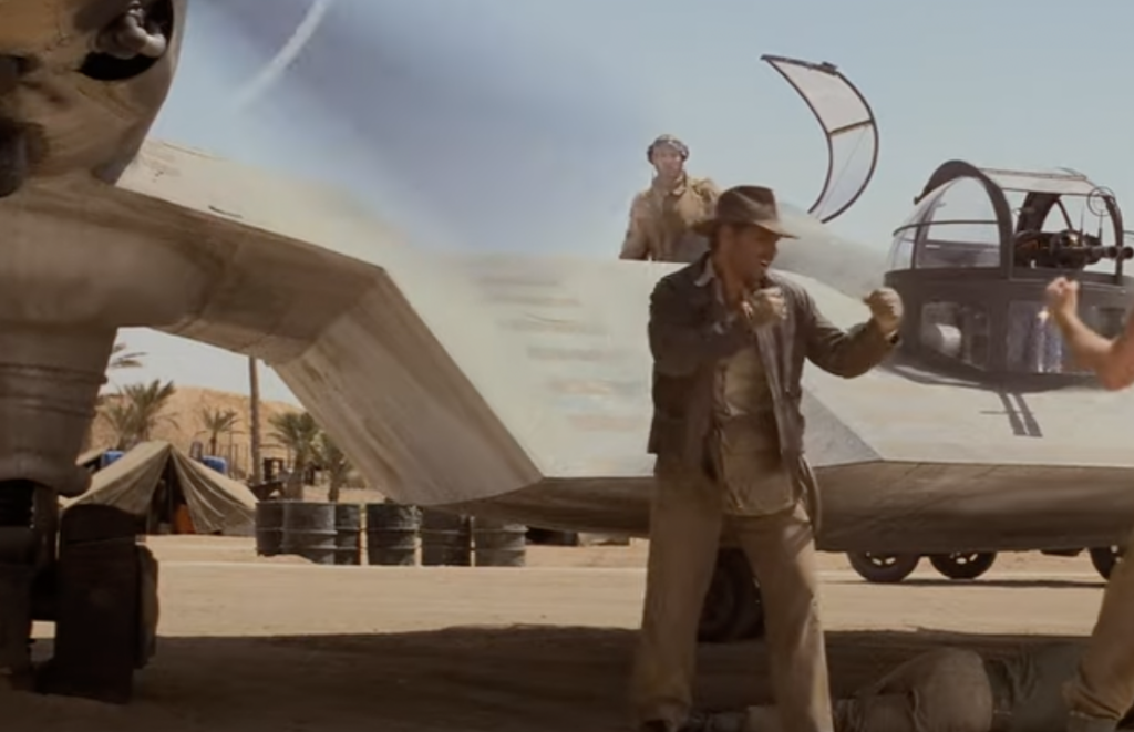 A scene from an action-adventure movie shows a man in a fedora and leather jacket preparing to fight another man near an airplane on a desert airfield. A person operates the plane's turret in the background amidst tents and palm trees.