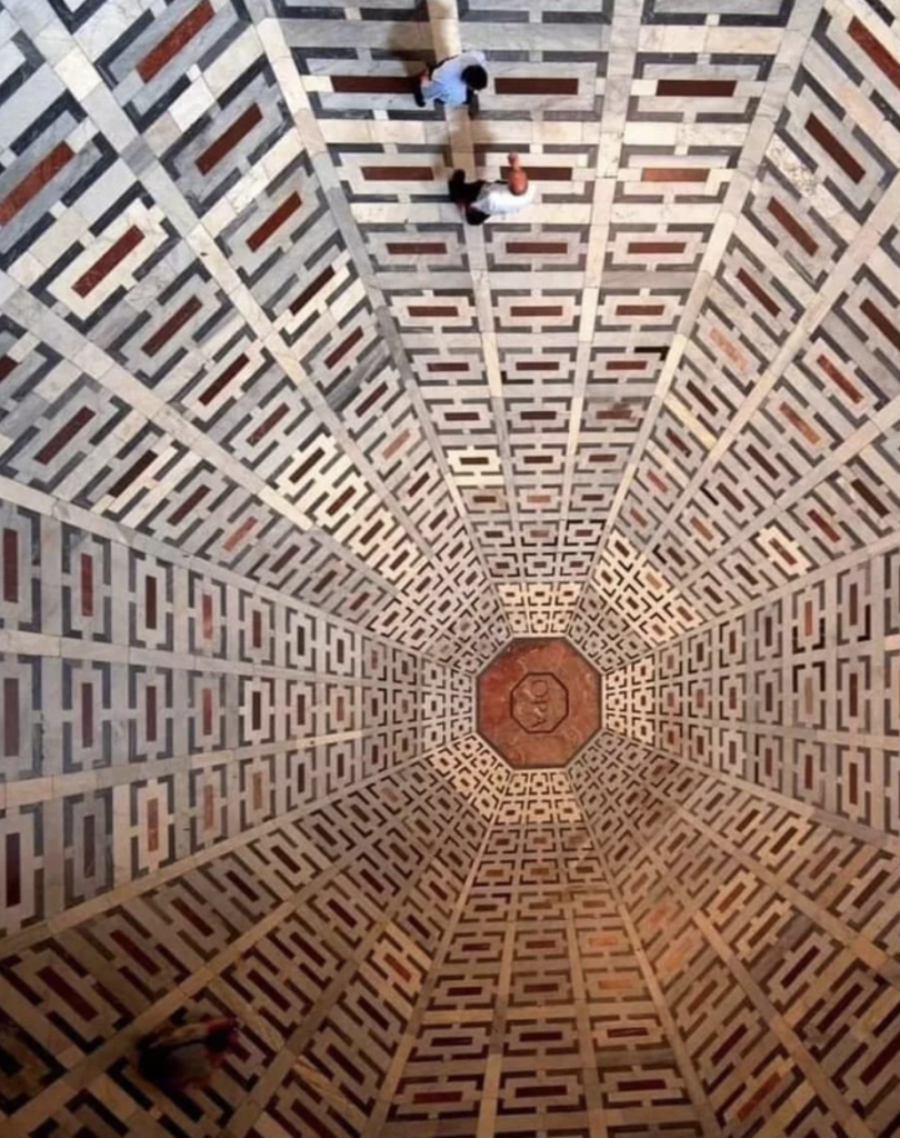 A geometric patterned tiled floor with intricate maze-like designs in shades of beige, brown, and red. Two people are walking across the floor, creating a sense of depth and perspective. The center features a prominent octagonal medallion.