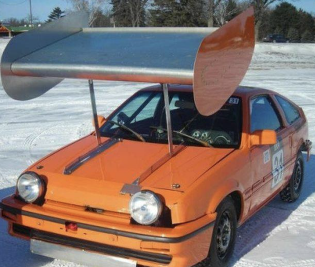 An orange car is equipped with an exceedingly large, homemade aluminum wing attached to its roof. The car appears to be in a snowy environment, with trees and clear sky in the background. The size of the wing is disproportionate to the size of the car.