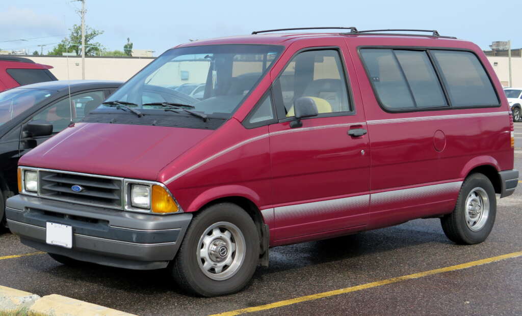 A maroon Ford minivan is parked in a parking lot. The vehicle has a boxy shape, grey lower paneling, and visible rubber bumper guards. The license plate is white, and there are other cars and a building in the background.