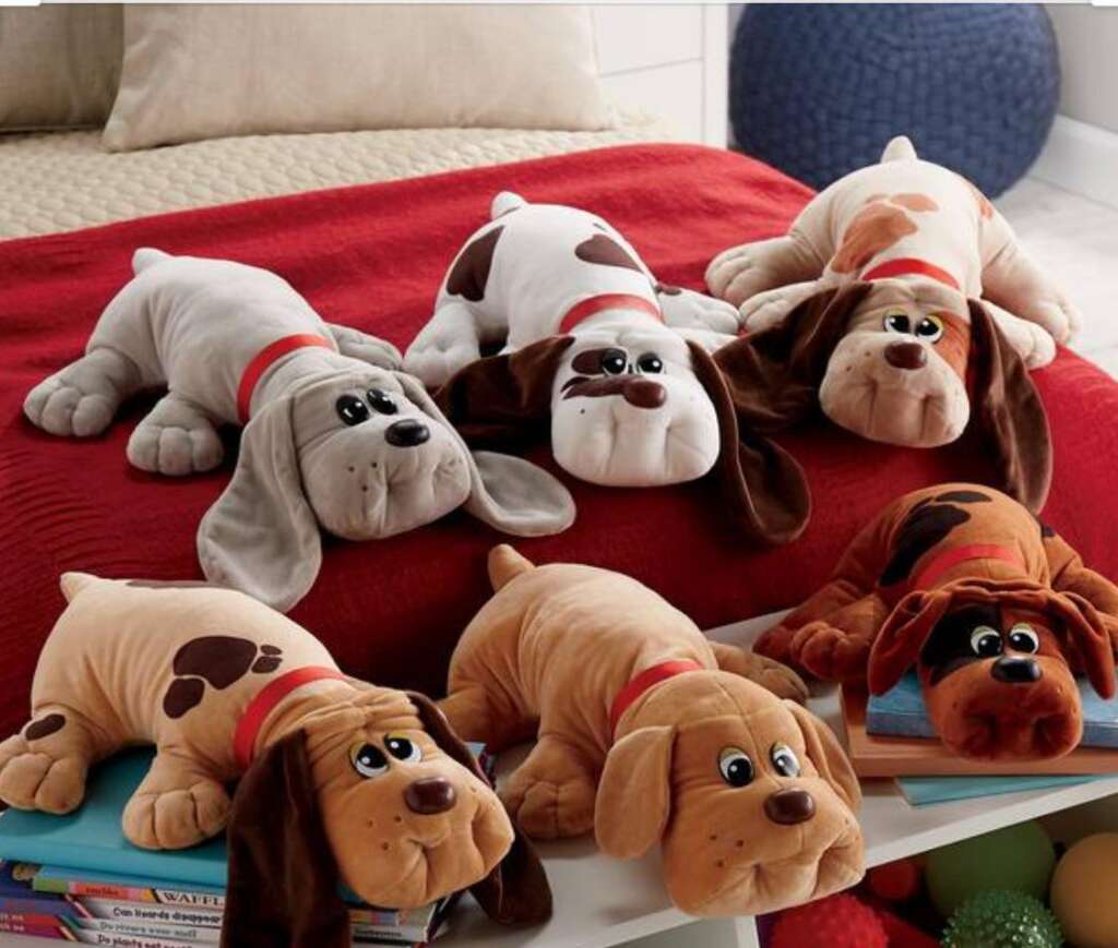 A group of six plush toy puppies with red collars are arranged on a red bedspread. They vary in colors, including white, brown, and tan, with some having spots. Each toy has large, expressive eyes and floppy ears, creating a cute and playful display.