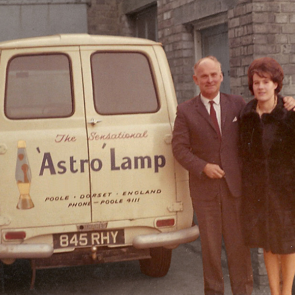 A man and woman stand beside a yellow van that has an advertisement for "The Sensational Astro Lamp" printed on its side. The man, dressed in a suit, has his arm around the woman, who is wearing a fur coat. The van’s license plate reads "845 RHY.