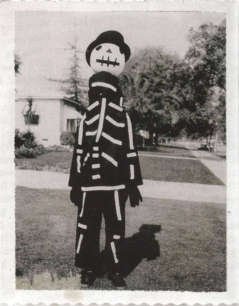 A person in a vintage skeleton costume stands outside on a sunny day. The costume features a black outfit with white bone designs and a round, pumpkin-like head with painted eyes, nose, and a mouth. Trees and a house are visible in the background.