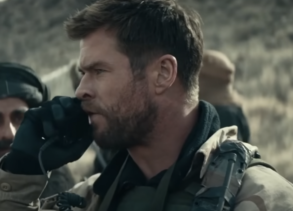 A man with short hair and a beard, wearing military gear and gloves, is speaking into a handheld radio. He appears to be in a rugged outdoor setting, with blurred figures and a hilly landscape in the background.