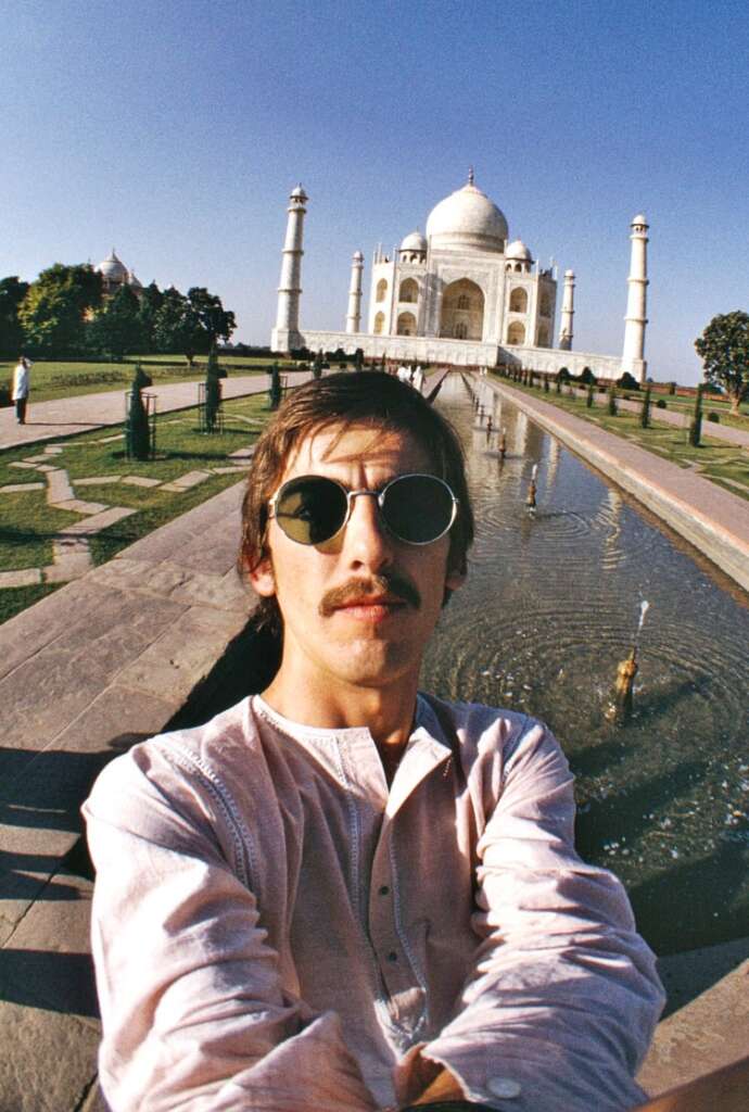 A person wearing sunglasses and a light pink shirt takes a selfie in front of the Taj Mahal. The iconic white marble mausoleum and its symmetrical minarets are visible in the background, with a reflective pool and surrounding gardens in the foreground.