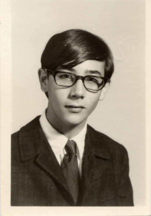 Black and white portrait of a young person with short hair, wearing glasses, a dark suit jacket, and a tie. The expression is neutral, and the background is plain. The photo appears vintage, likely from the mid-20th century.