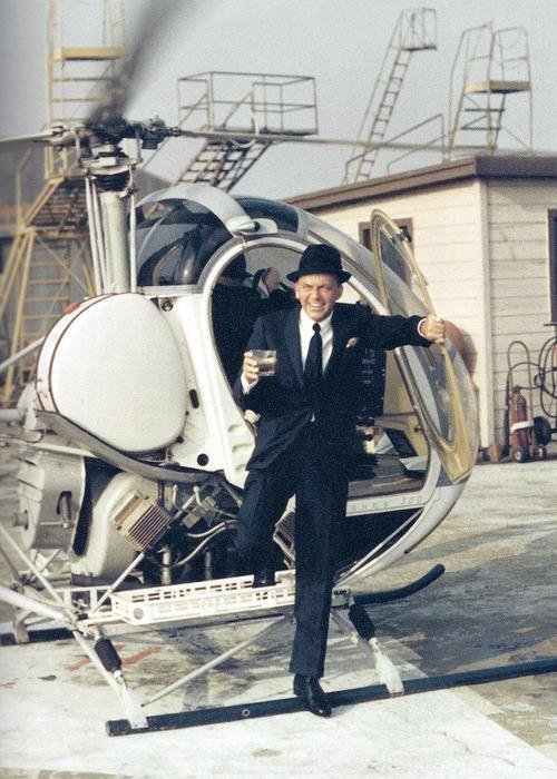 A man dressed in a suit and tie steps out of a helicopter, holding a drink in one hand and the helicopter door in the other. He is smiling, and the helicopter is parked on a rooftop with various industrial structures in the background.